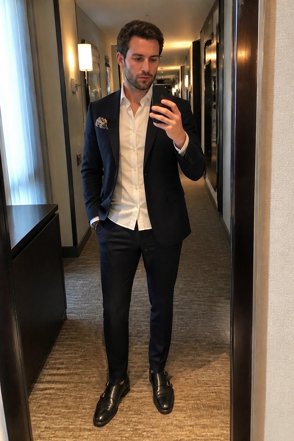 Man in navy suit taking mirror selfie with phone in hotel hallway