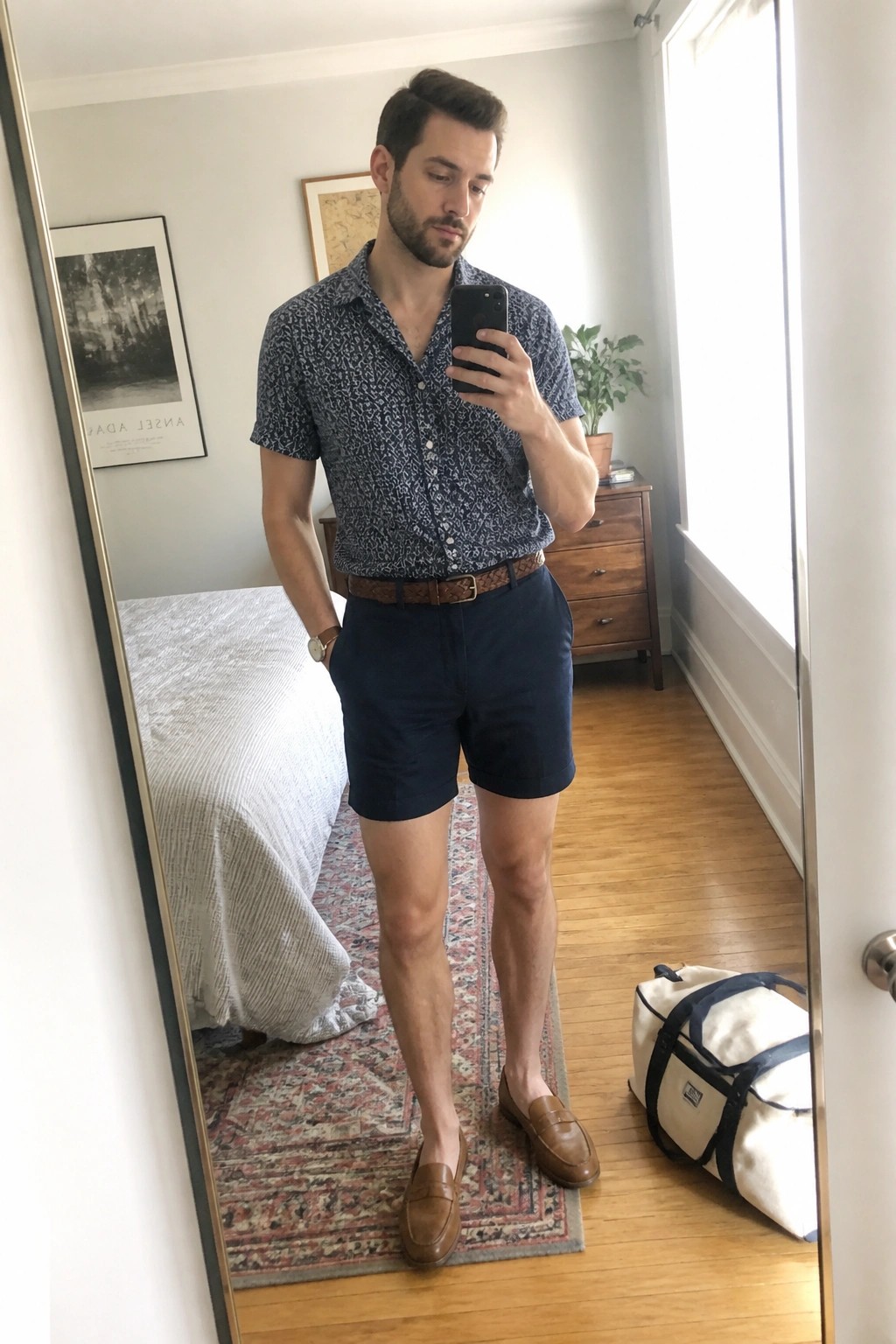 Man in patterned shirt and navy shorts takes mirror selfie in bedroom