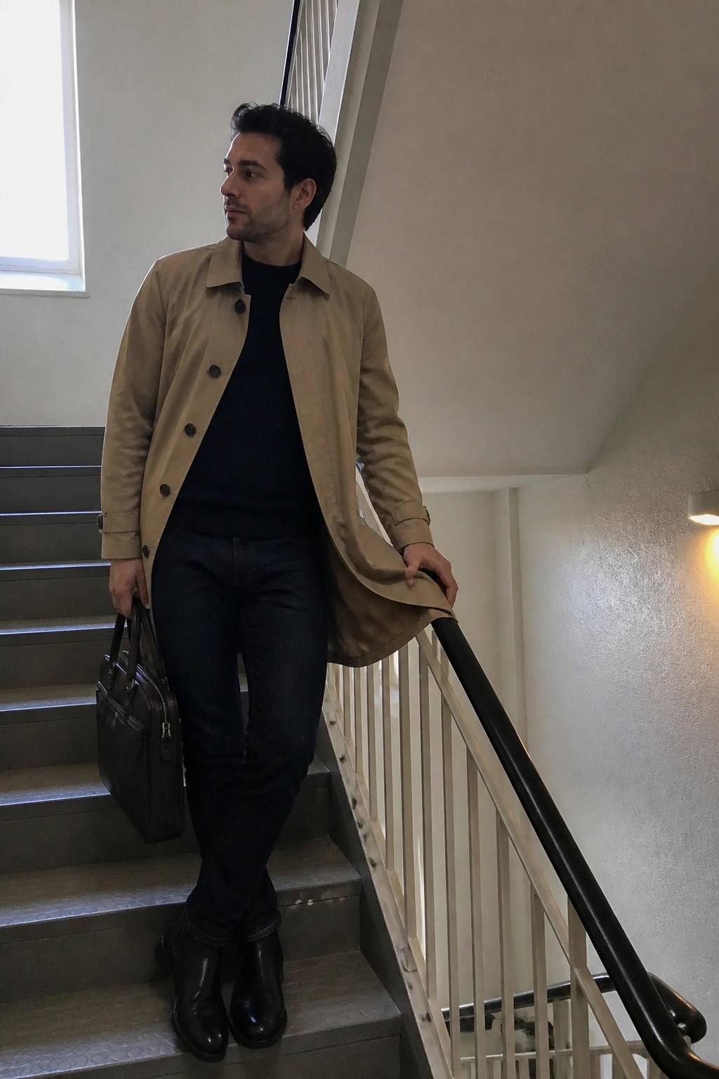 Man in tan coat over black sweater holding briefcase on stairs
