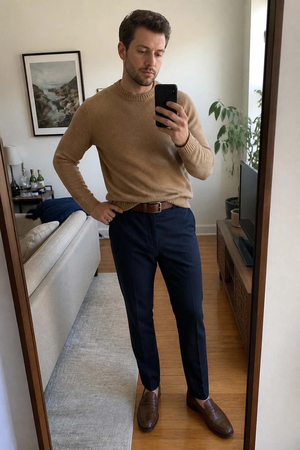 Man in tan sweater and navy pants taking mirror selfie in living room