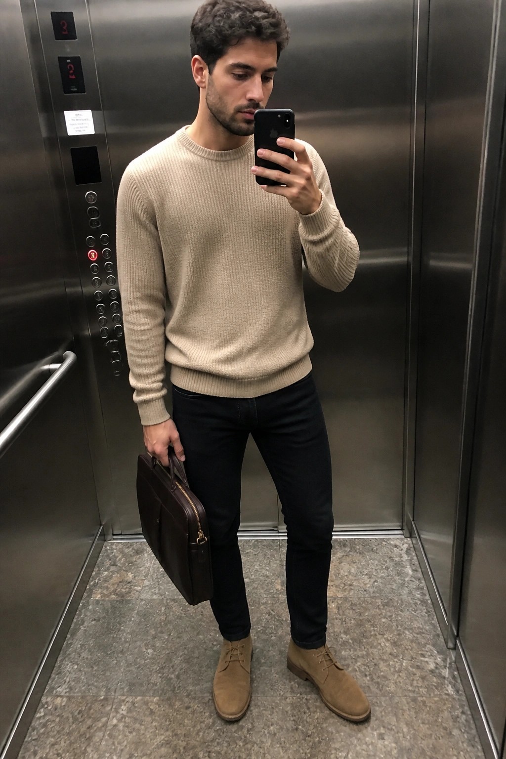 Man in beige sweater holding phone and briefcase inside elevator.