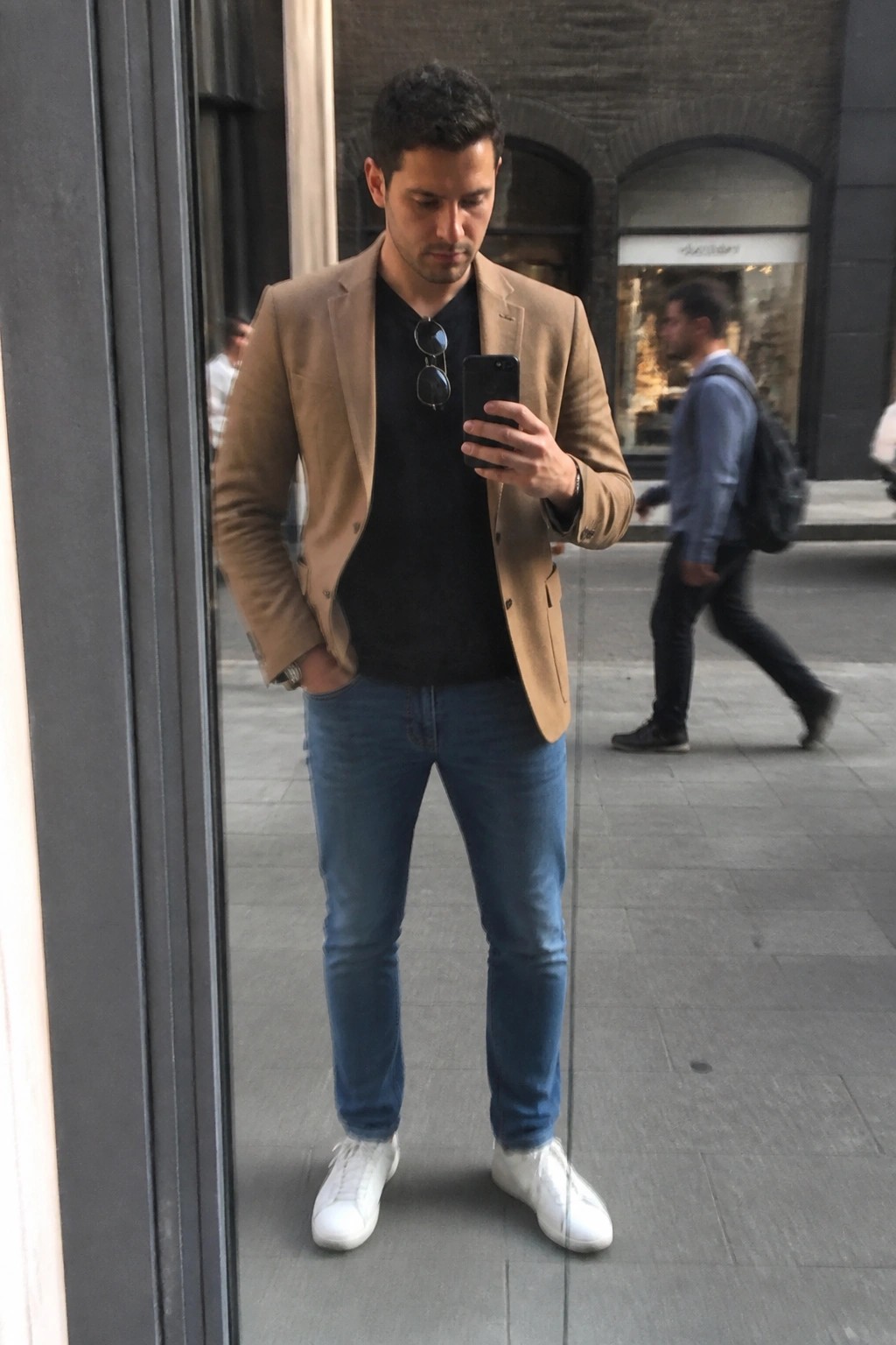 Man in tan blazer and jeans taking mirror selfie on city street