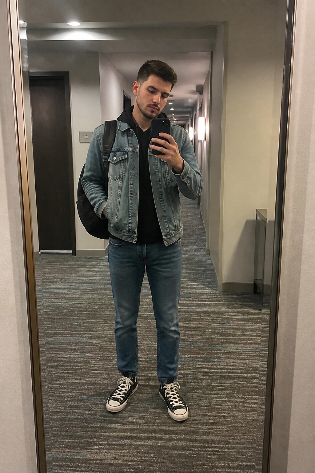 Man in denim jacket and jeans taking mirror selfie in carpeted hallway with backpack