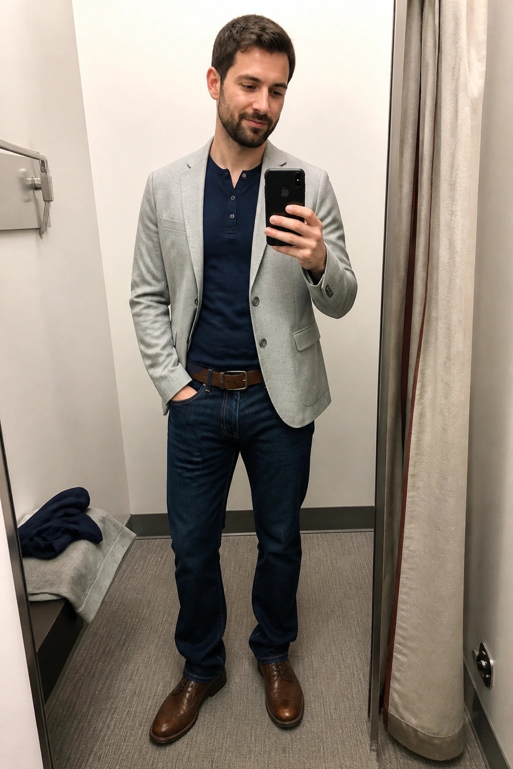 Man in gray blazer and jeans taking mirror selfie with iPhone in dressing room.