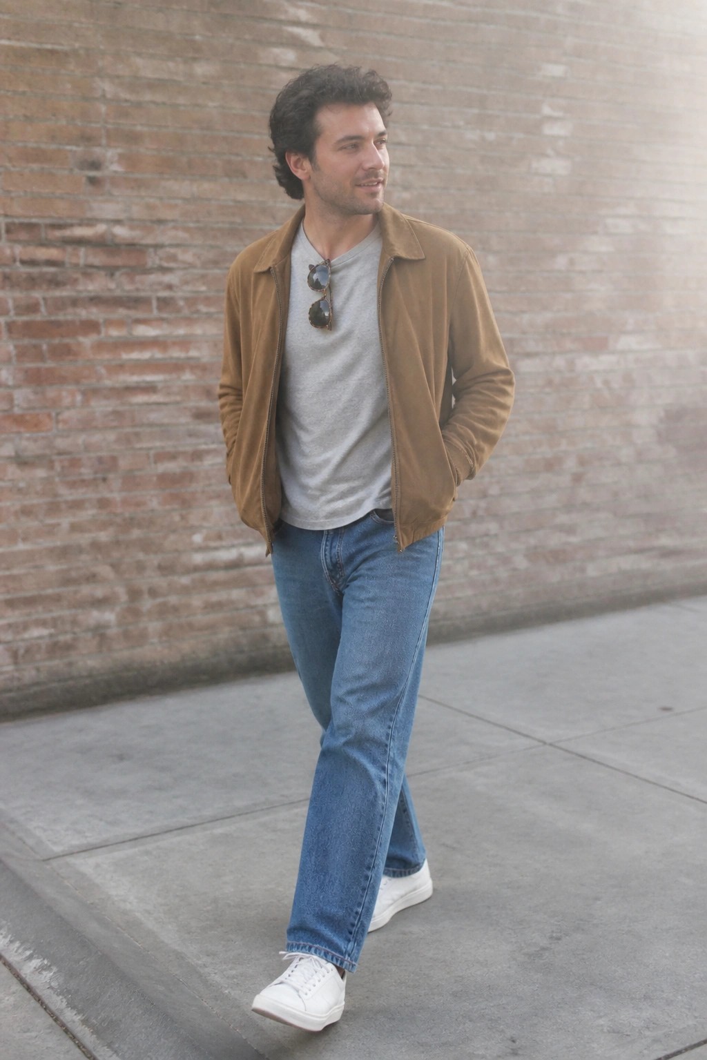 Man in brown suede jacket over gray t-shirt with blue jeans and white sneakers by brick wall
