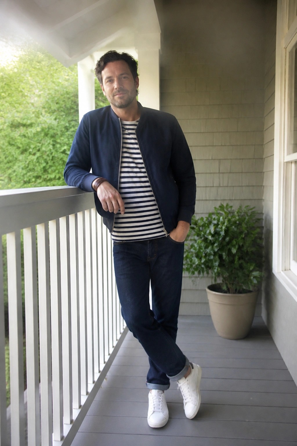 Man wearing navy bomber jacket over navy and white striped t-shirt with dark blue jeans cuffed at the ankles and white sneakers standing on a porch.