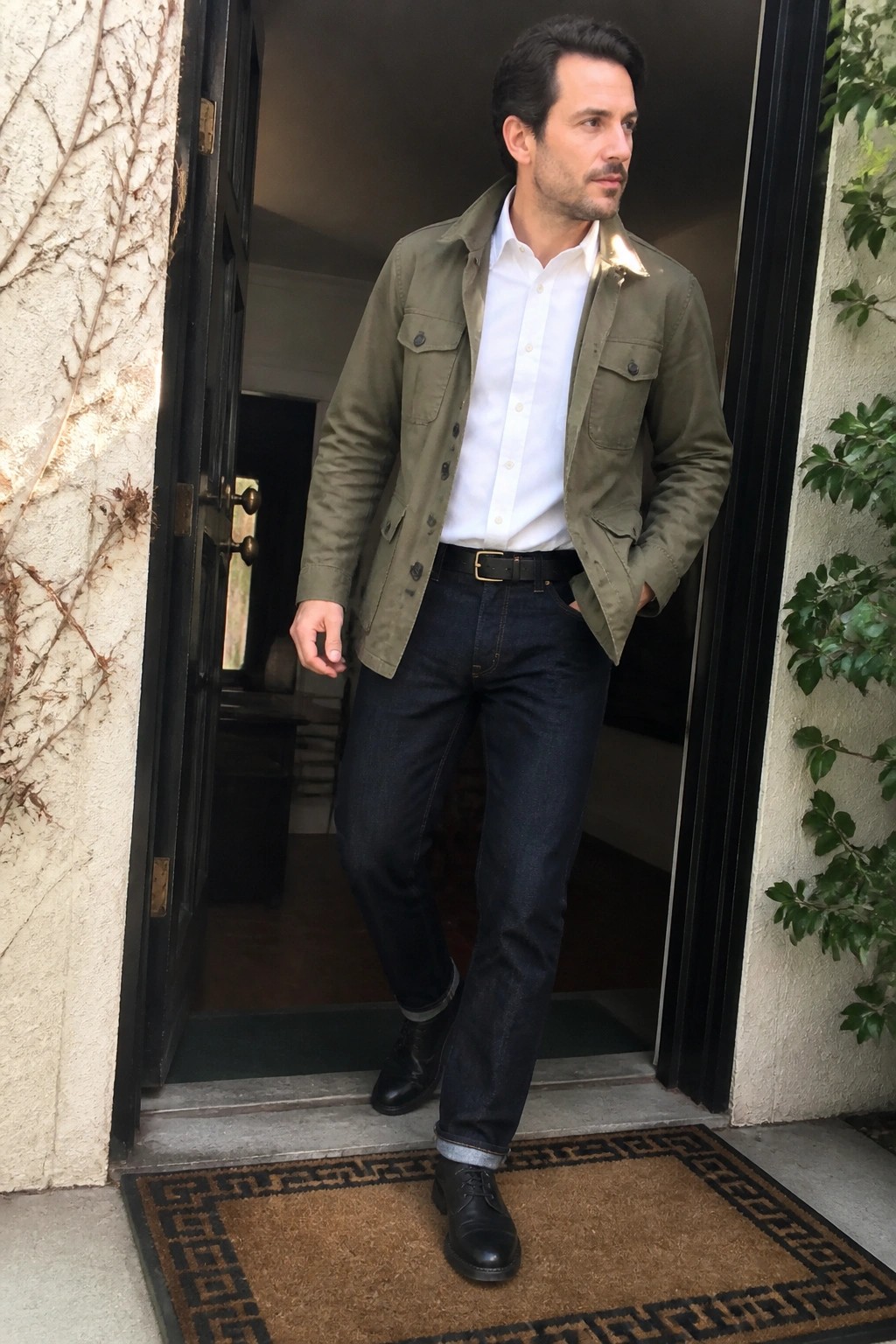 Man in olive jacket over white shirt with dark jeans and black shoes in doorway