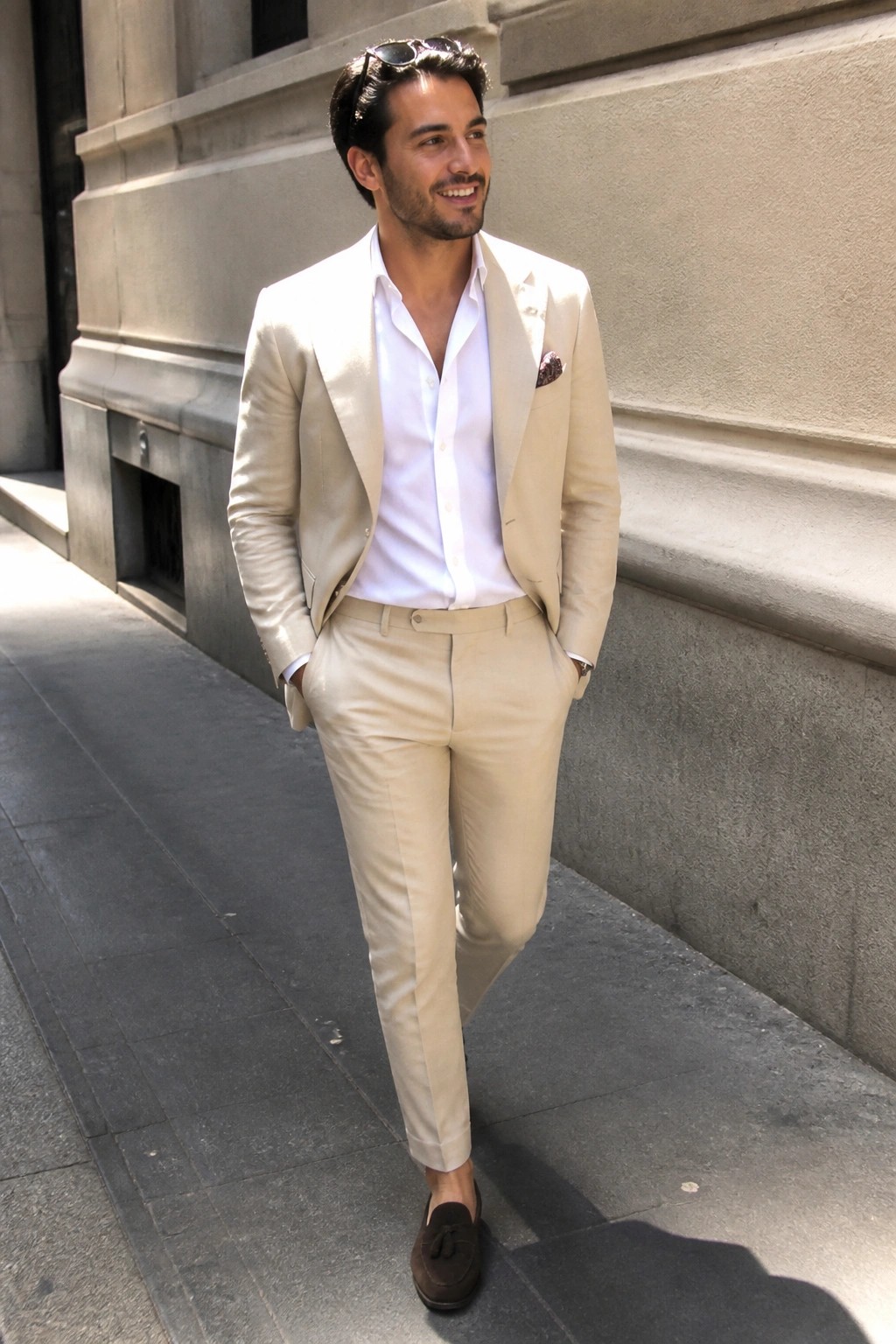 Man in beige suit and white shirt stands on sidewalk by stone building