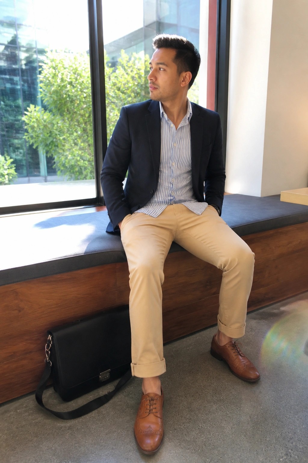 Man in navy blazer and khaki pants sits by window with black bag
