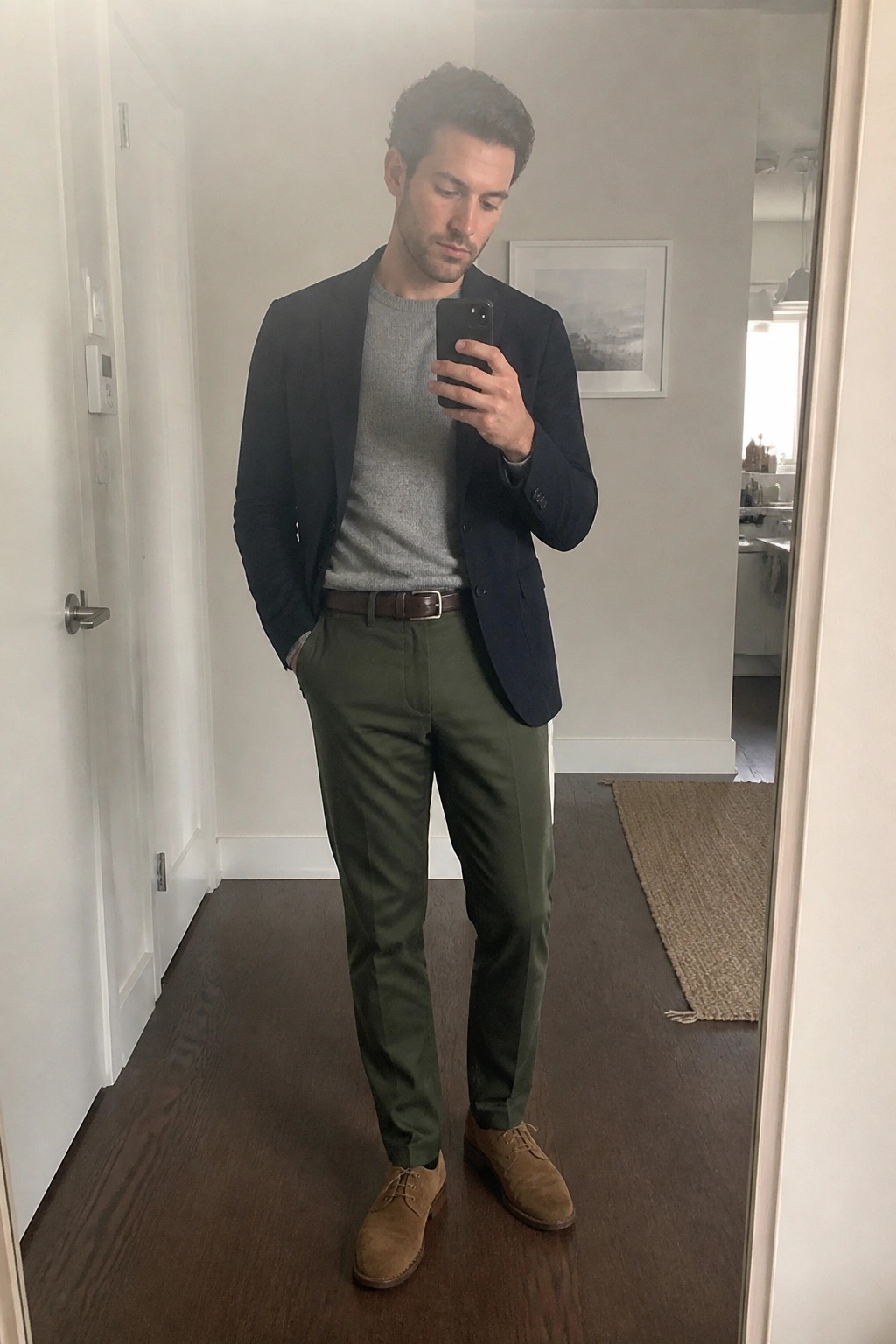 Man in navy blazer over gray sweater with olive pants takes mirror selfie.