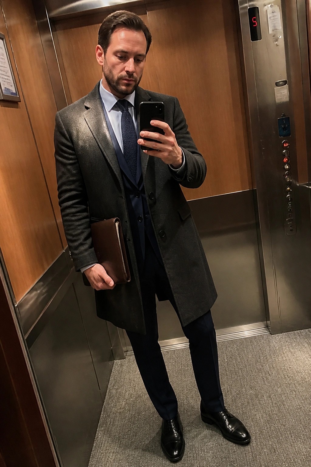 Man in gray overcoat over navy three piece suit with blue shirt patterned tie black dress shoes and brown leather portfolio standing in elevator taking mirror selfie.