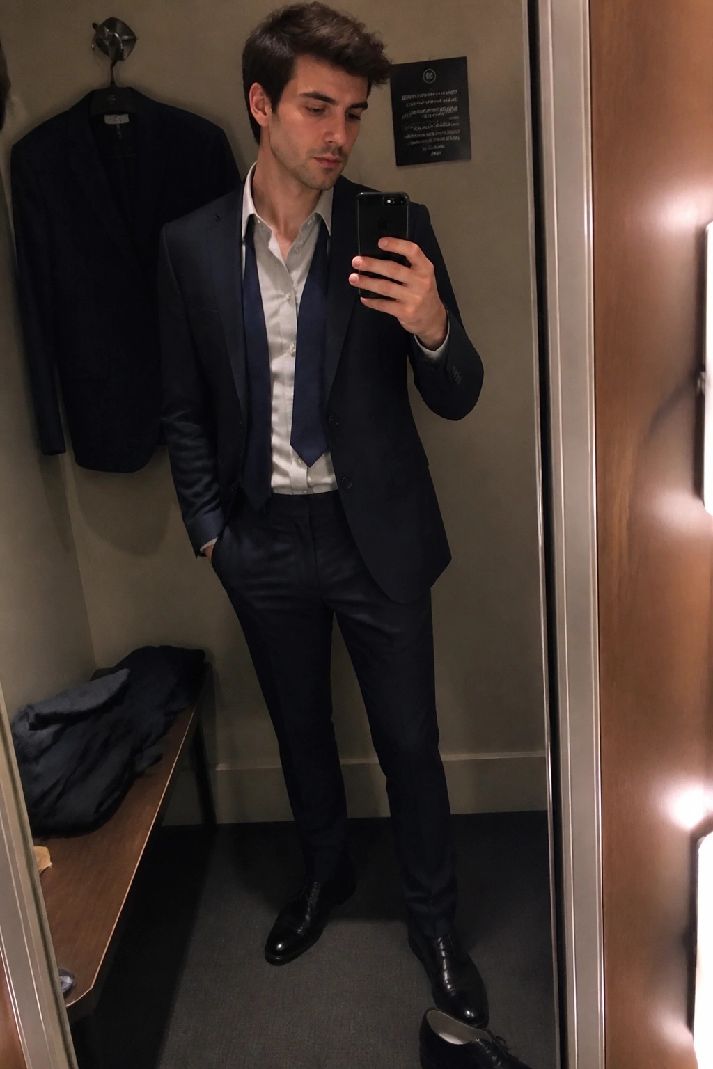 Man in navy suit takes mirror selfie in fitting room with phone