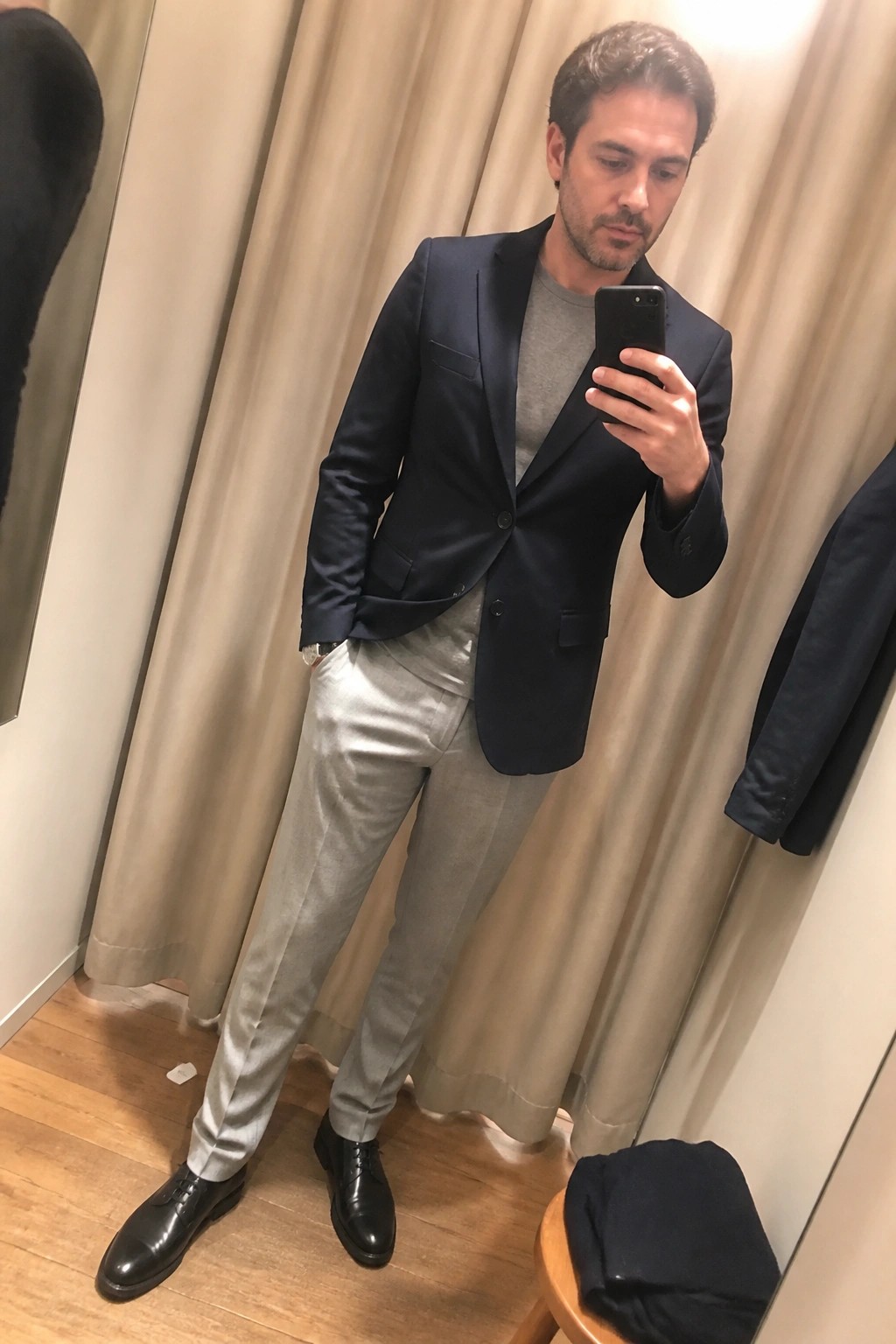 Man in navy blazer and gray pants taking mirror selfie in fitting room