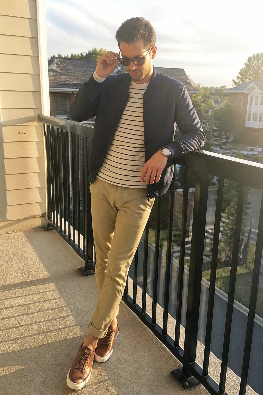 Man in striped shirt and bomber jacket adjusts sunglasses on sunny balcony.