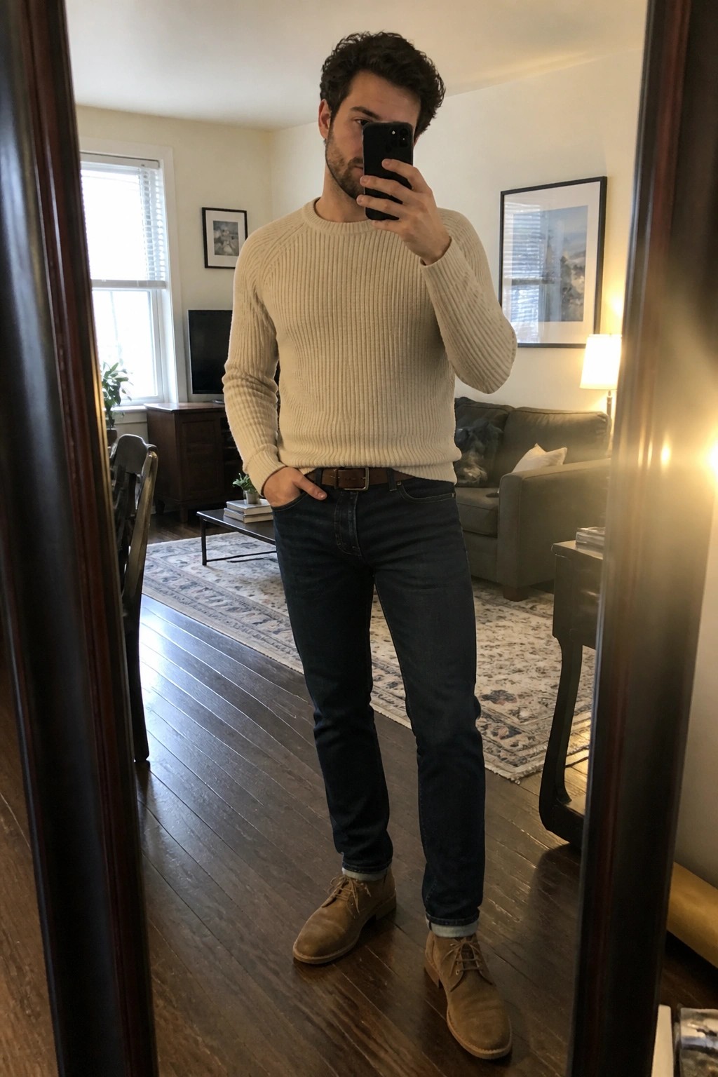 Man in beige sweater and dark jeans taking mirror selfie in living room