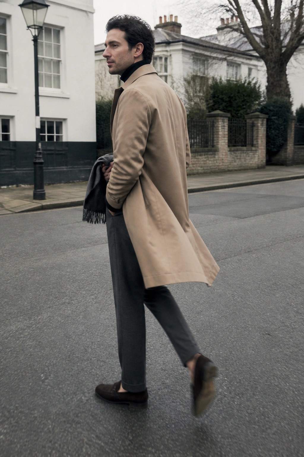 Man in beige trench coat walks on street holding fringed gray scarf