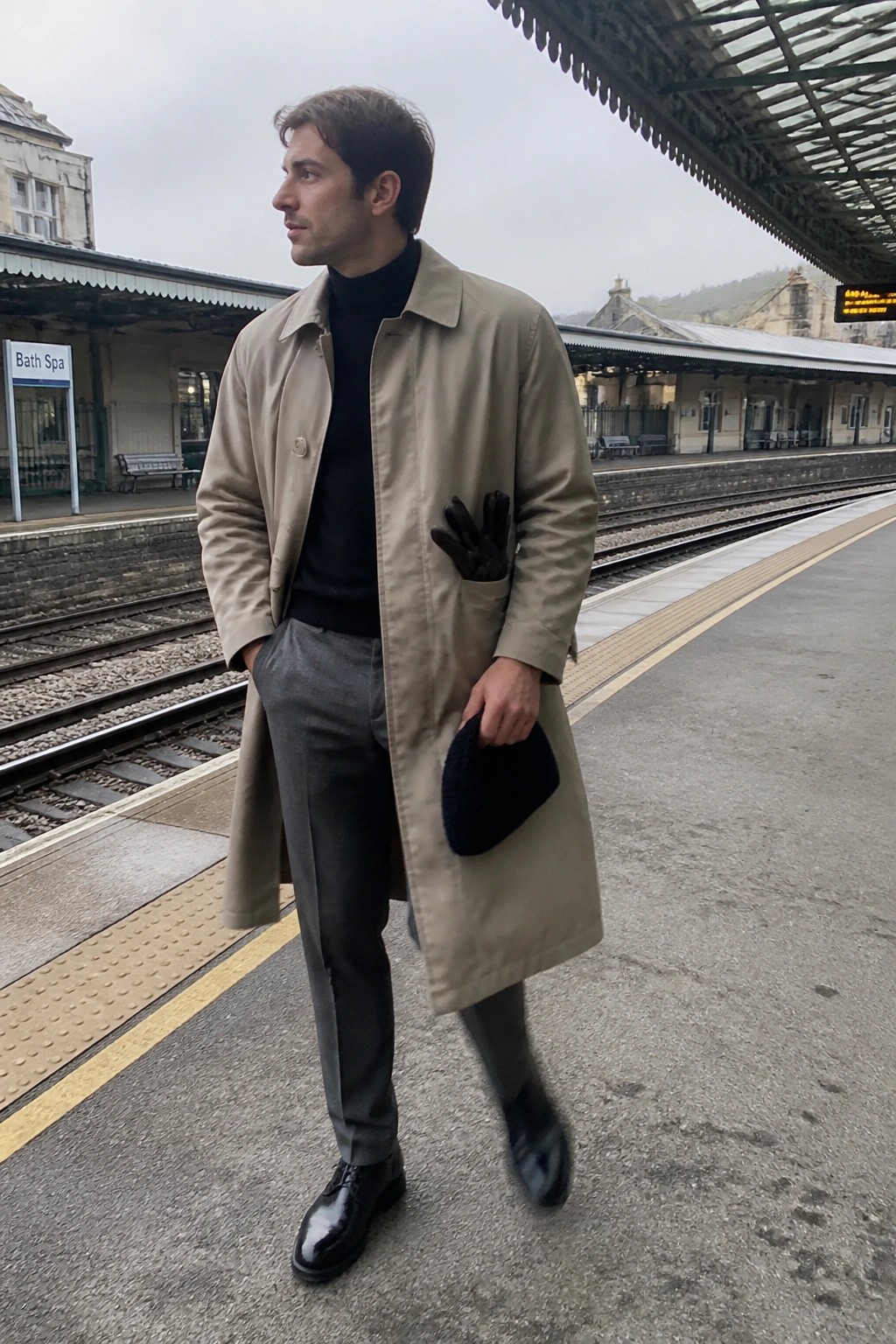Man in beige trench coat holding black hat at Bath Spa station platform