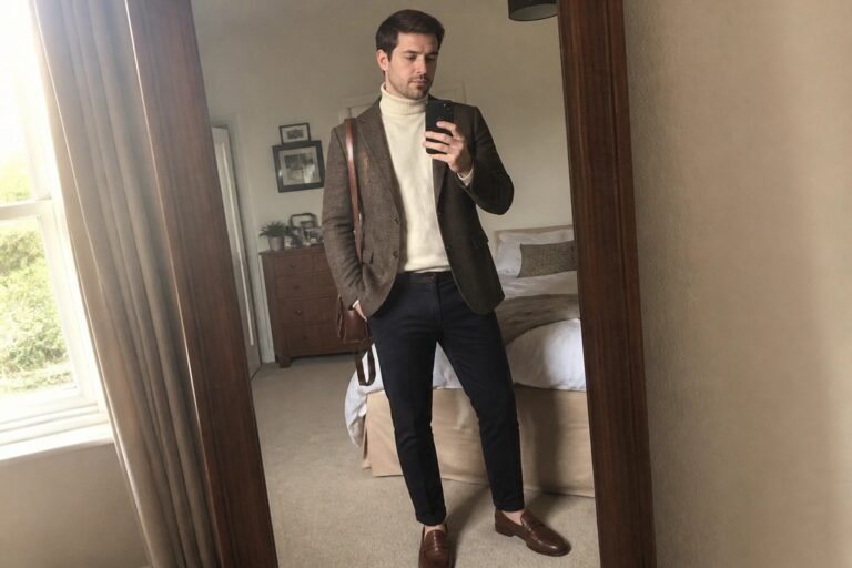 Man in brown tweed blazer over white turtleneck taking bedroom mirror selfie with leather bag