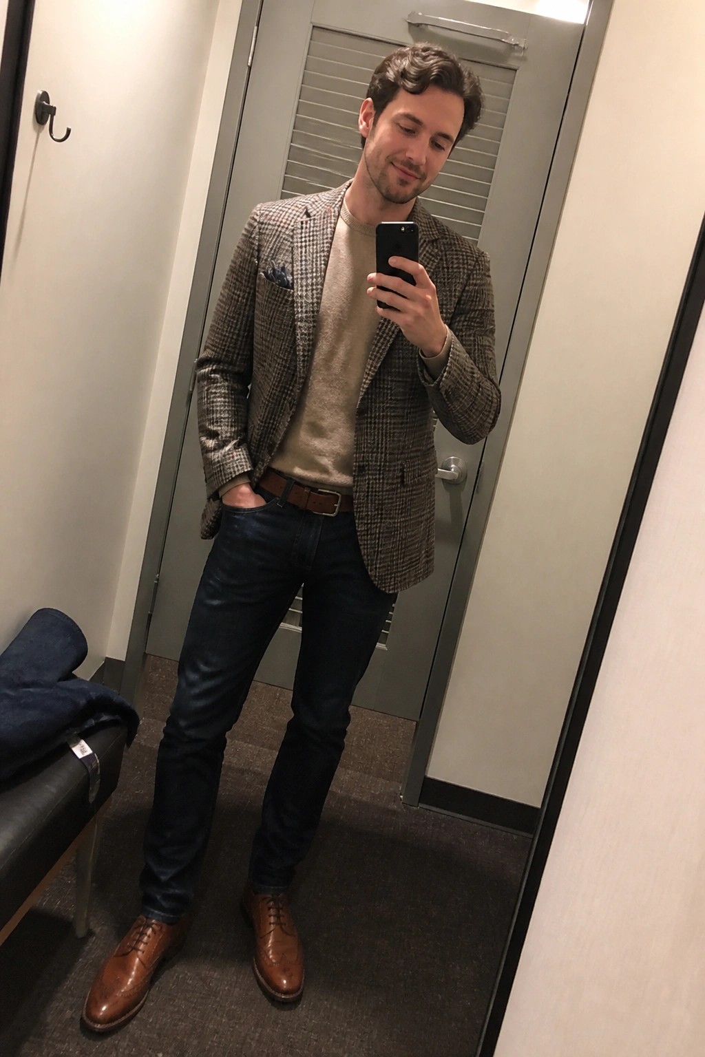 Man in houndstooth blazer over beige sweater takes mirror selfie in fitting room.