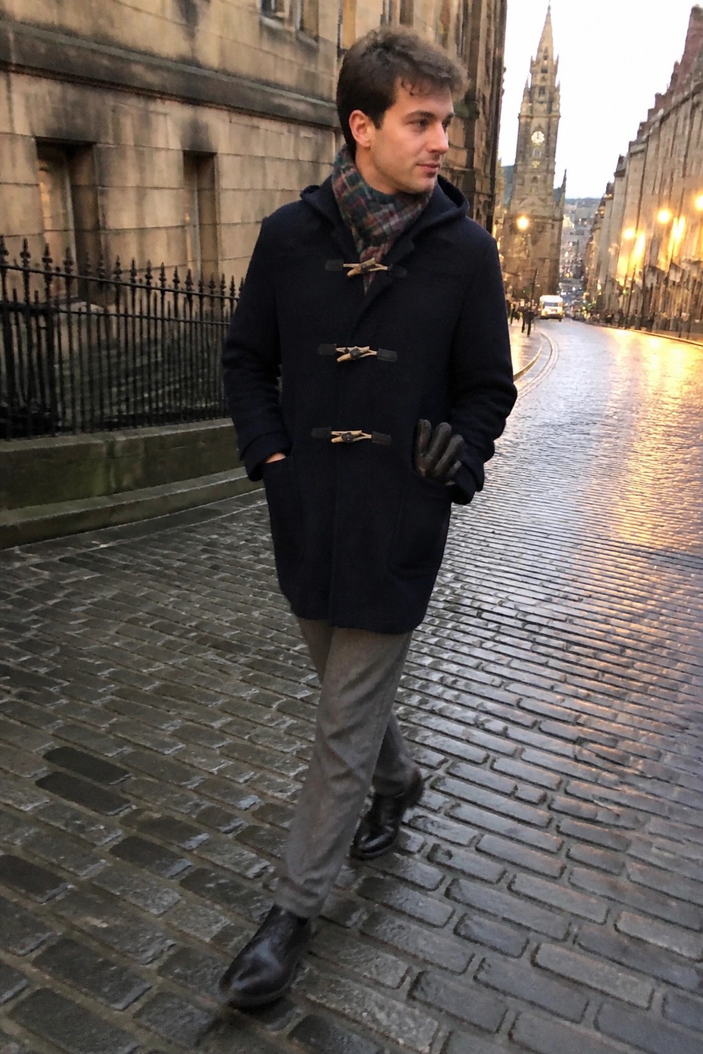 Man in navy duffle coat and plaid scarf walks on wet cobblestone street at dusk