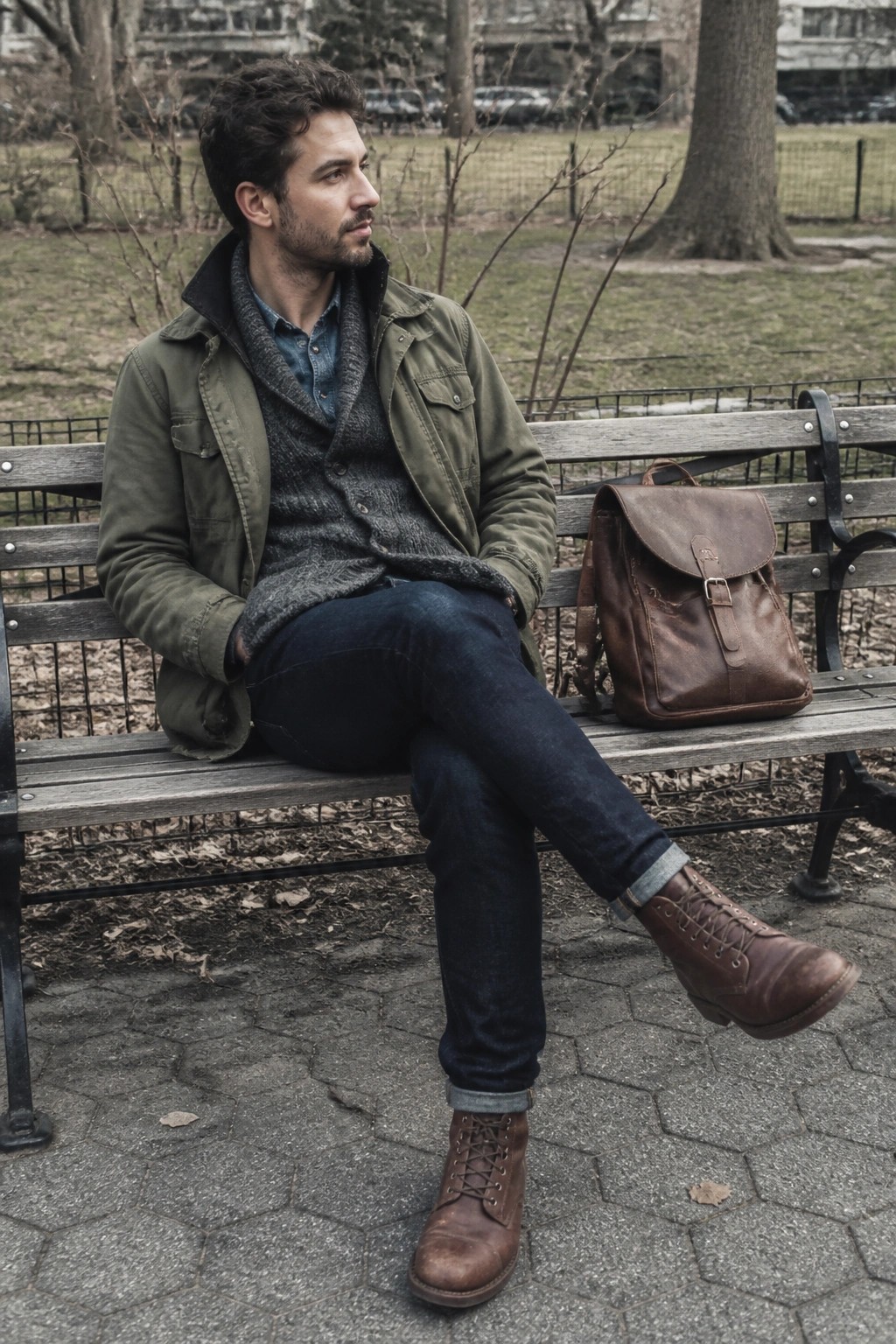 Man in green jacket seated on park bench beside brown leather backpack