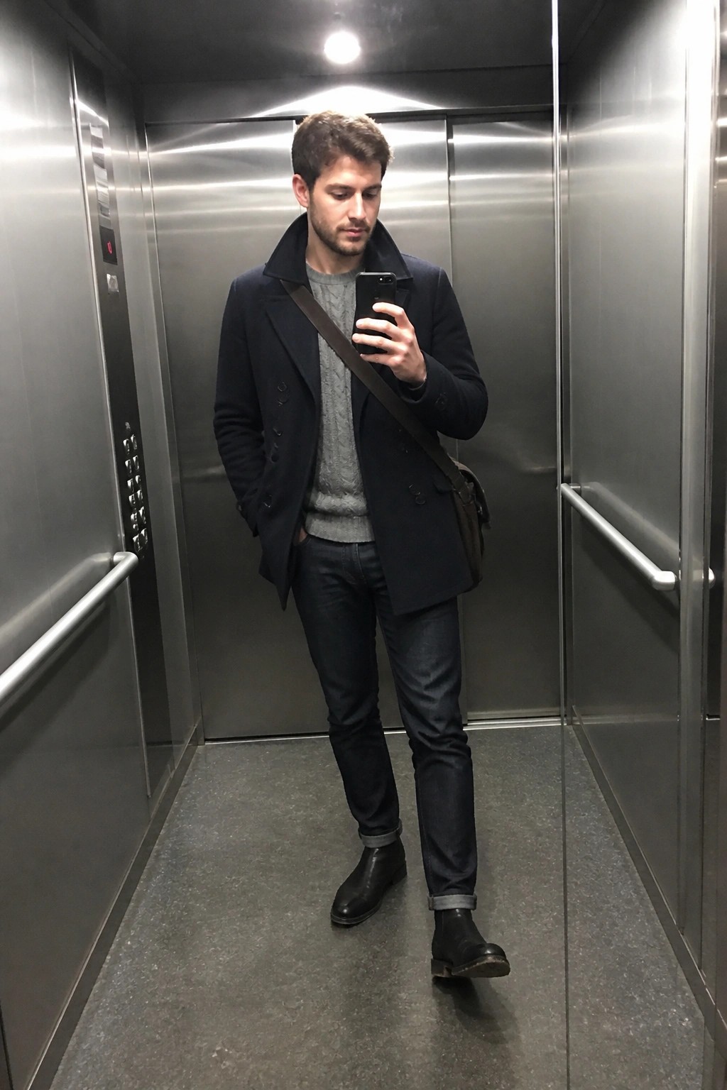 Man in navy peacoat taking selfie with phone in metallic elevator.