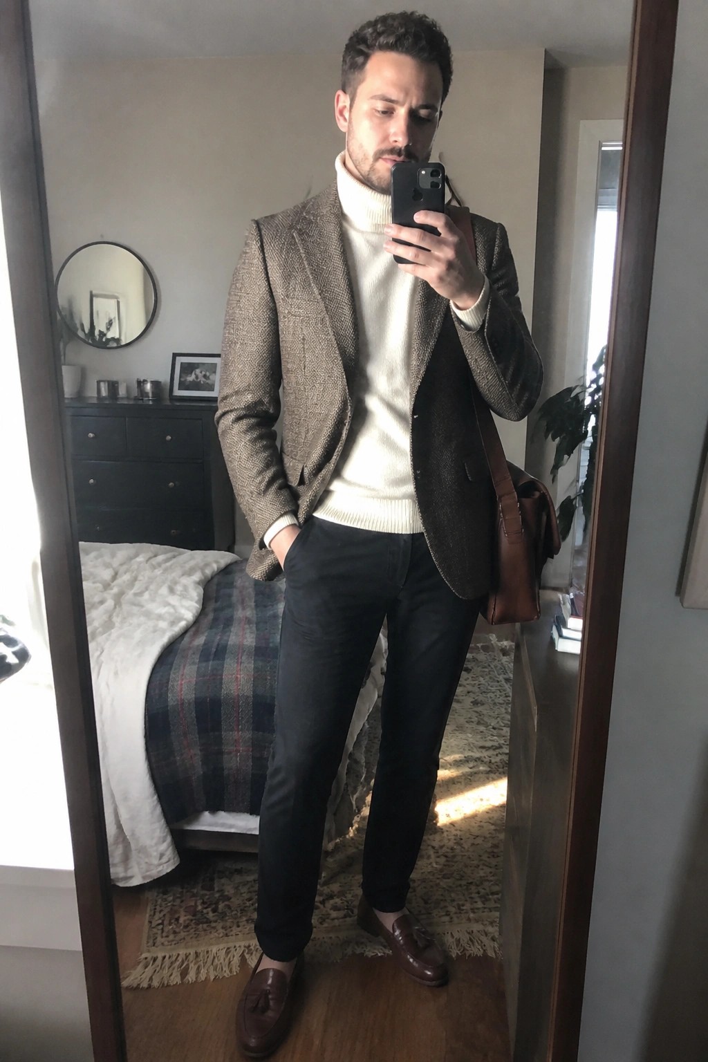 Man in brown tweed blazer over white turtleneck taking bedroom mirror selfie with leather bag