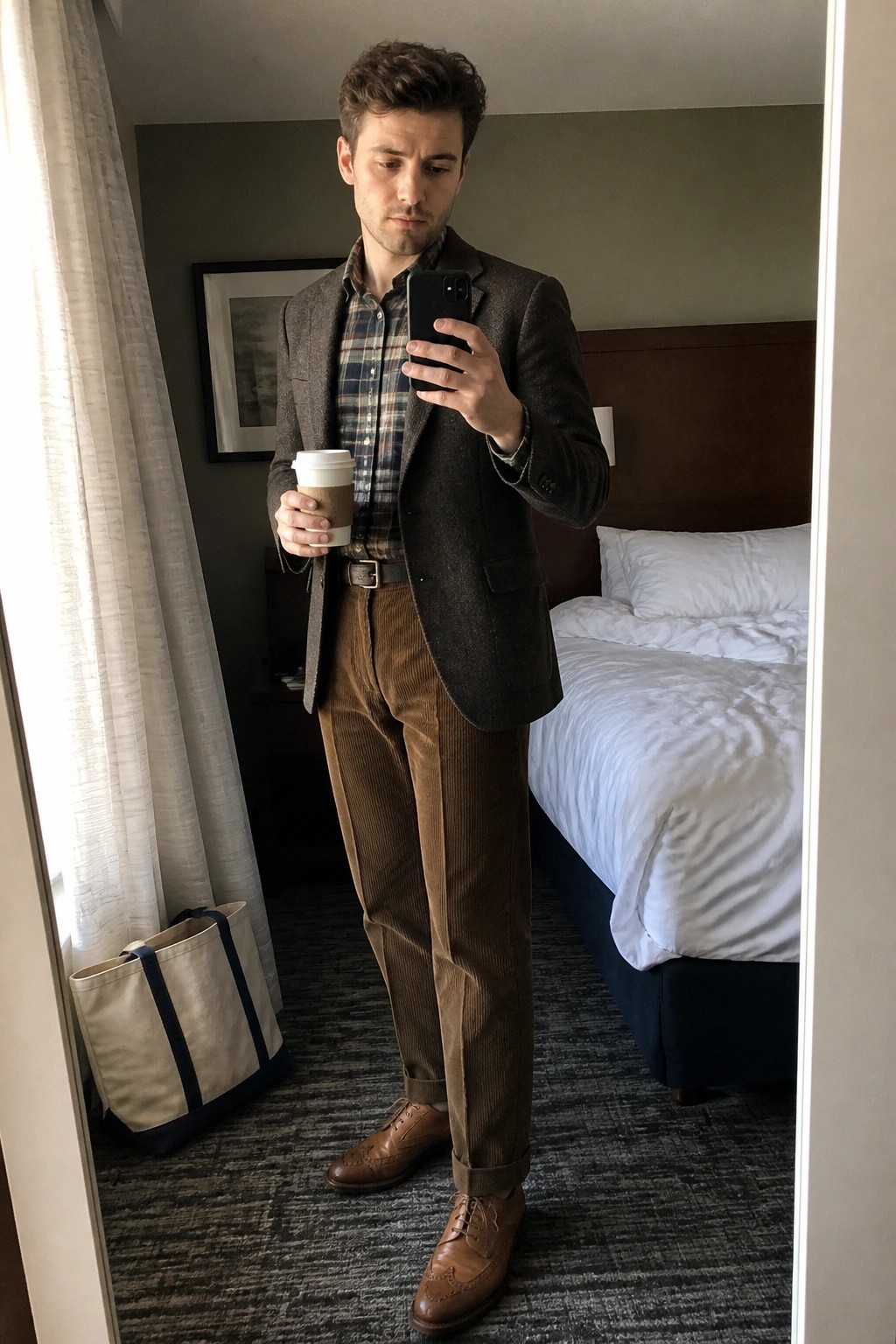 Man in brown blazer and corduroy pants taking mirror selfie while holding coffee