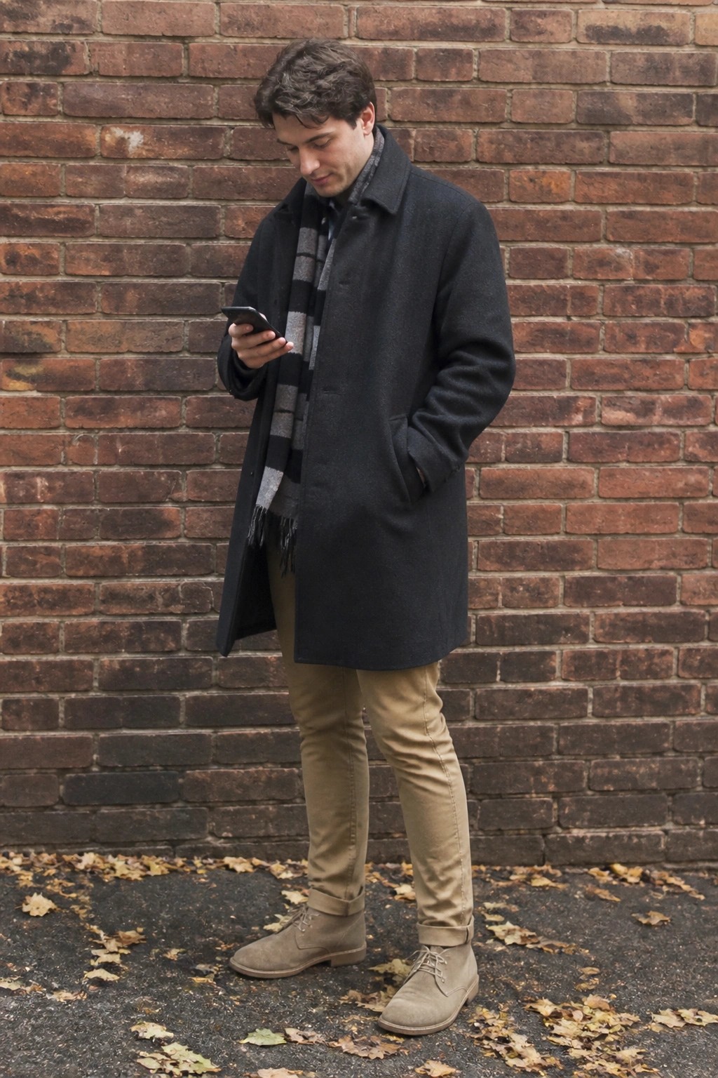 Man in dark coat and striped scarf checks phone by brick wall with autumn leaves