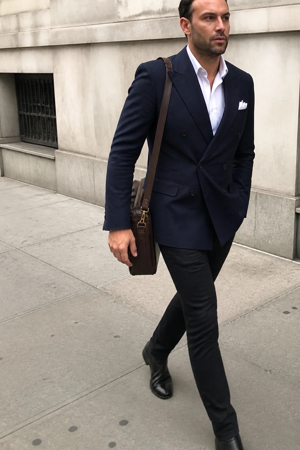 Man in navy double-breasted suit carrying brown leather bag on city sidewalk