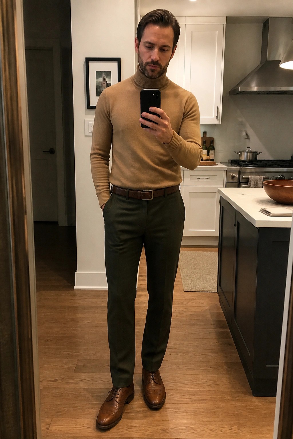 Man in tan turtleneck and olive pants taking mirror selfie in kitchen