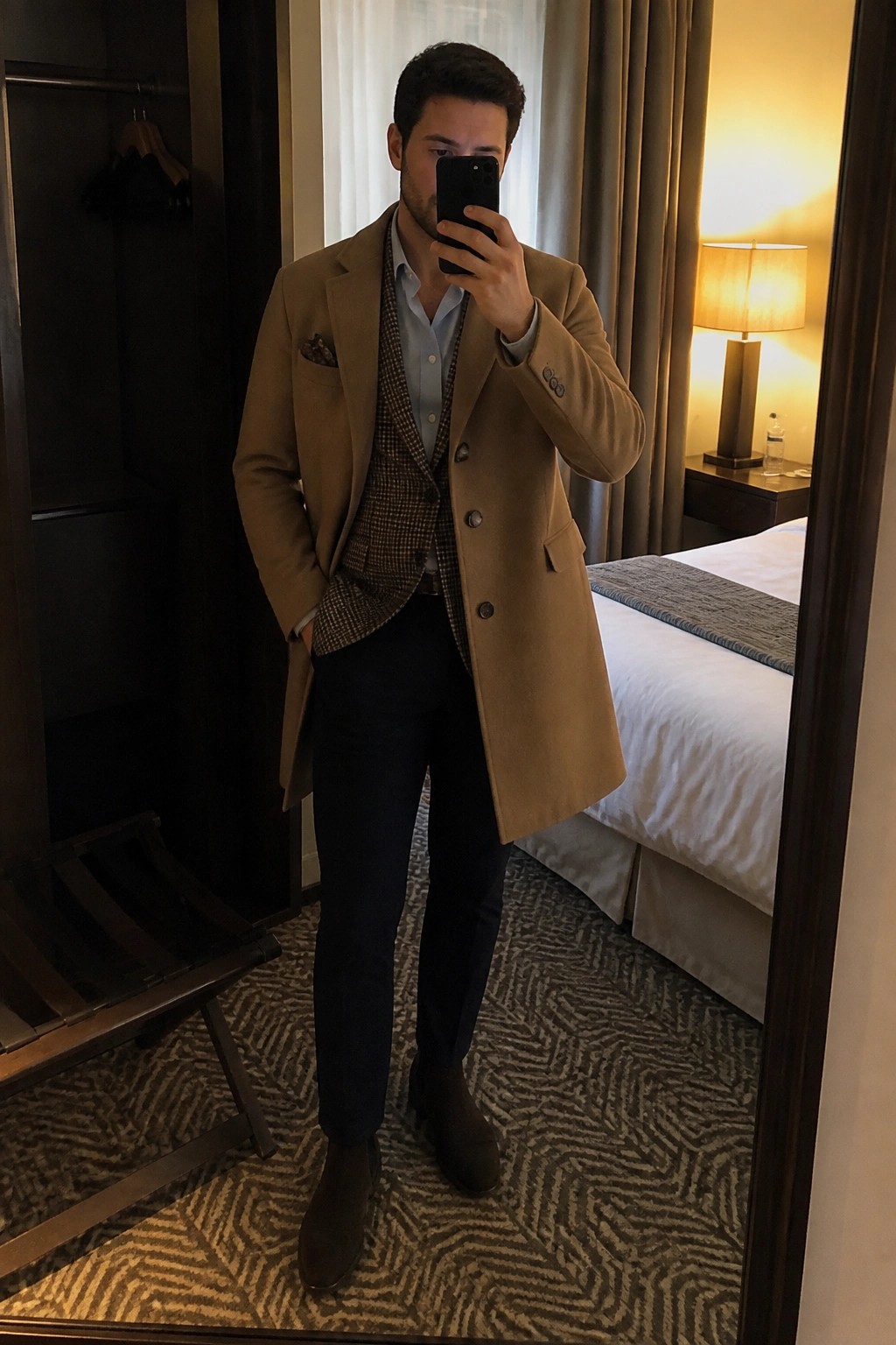 Man in tan overcoat and houndstooth vest taking mirror selfie in hotel room