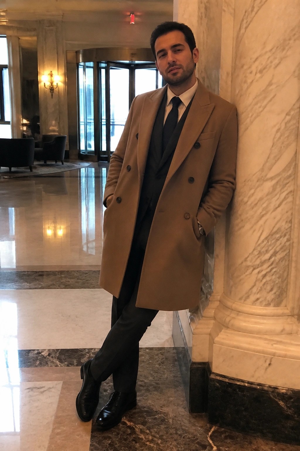 Man in tan overcoat leaning against marble pillar in elegant lobby
