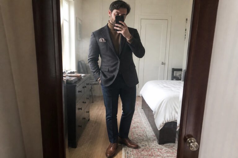 Man in gray blazer over tan turtleneck taking mirror selfie in bedroom