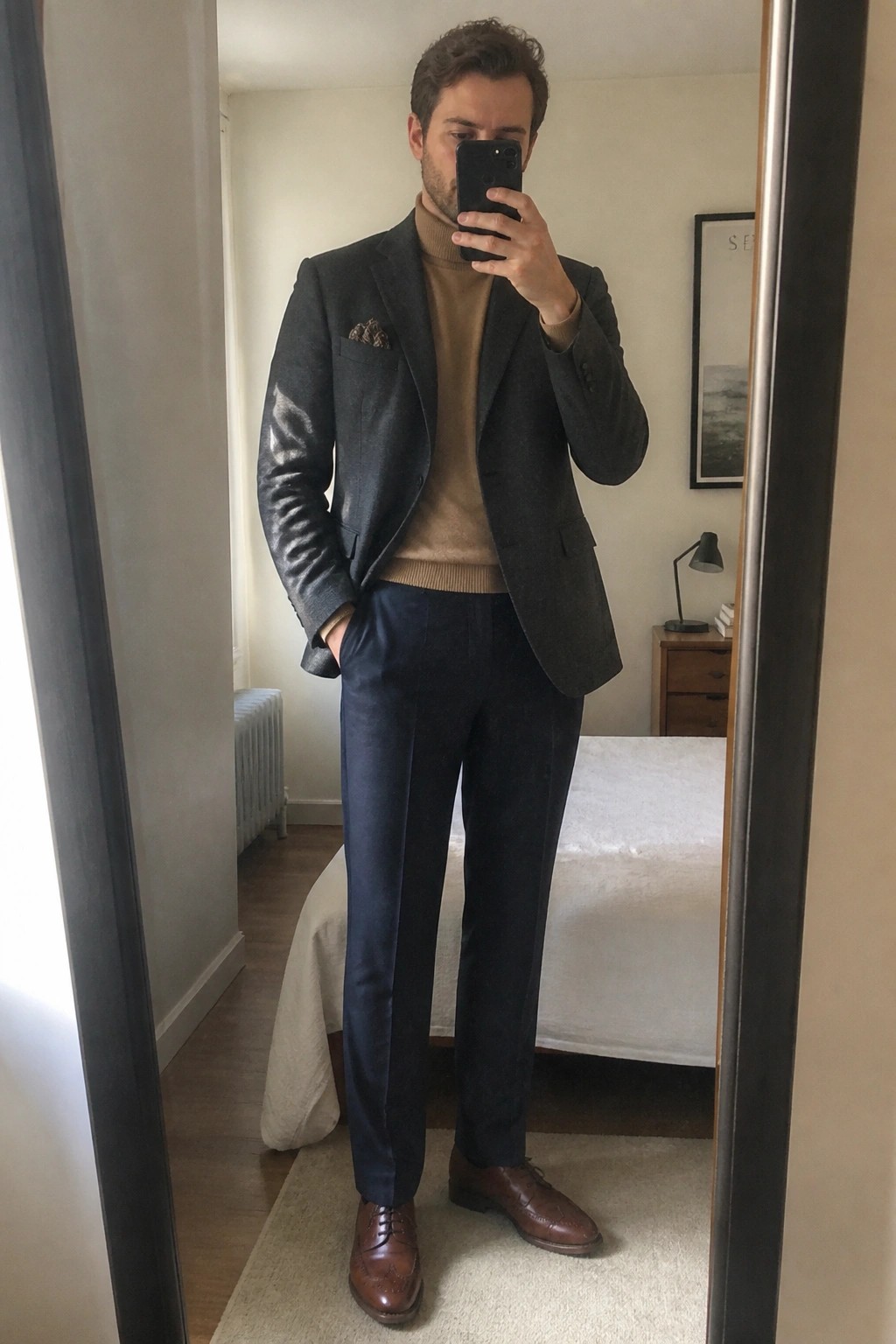 Man in gray blazer over tan turtleneck taking mirror selfie in bedroom