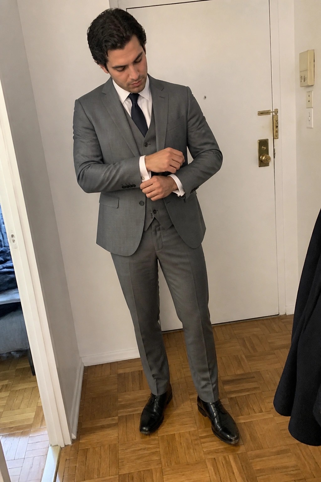 Man in gray three-piece suit adjusting cufflinks on hardwood floor indoors.