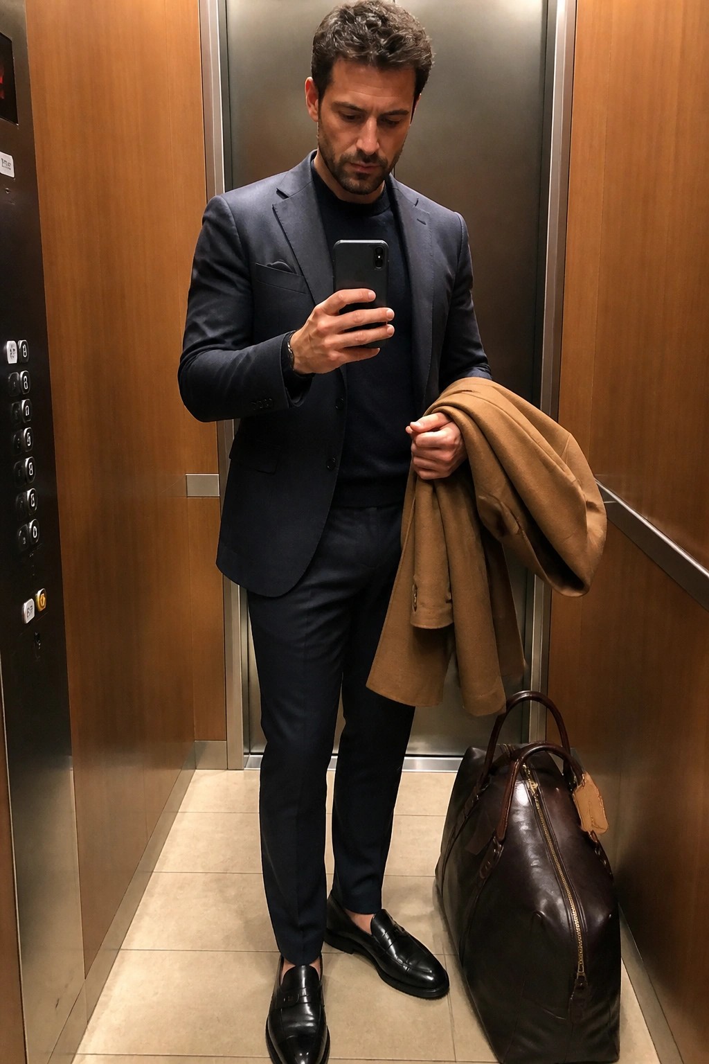 Man in navy suit checks phone while holding tan coat beside leather bag in elevator