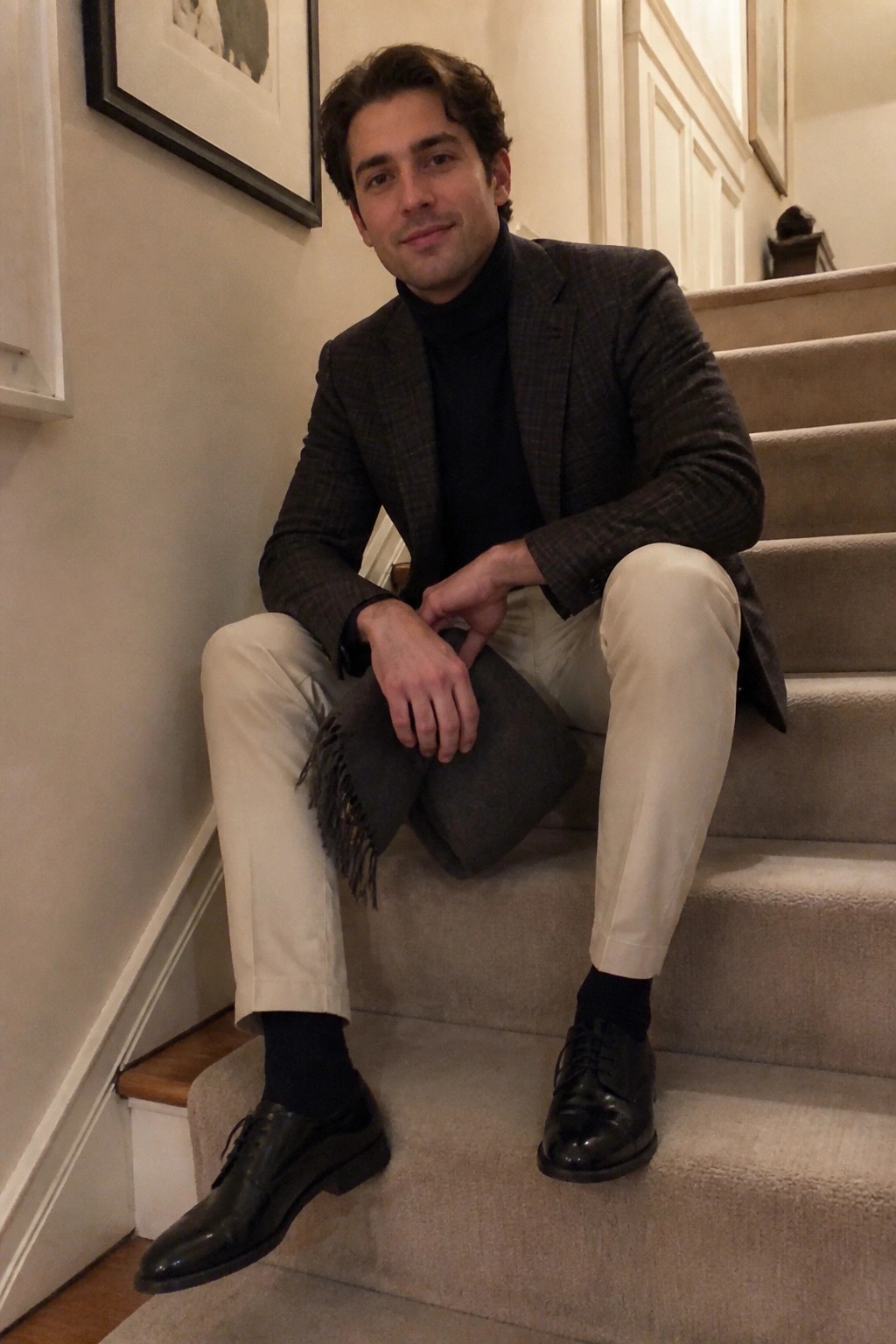 Man in plaid blazer and turtleneck seated on carpeted staircase holding gray scarf