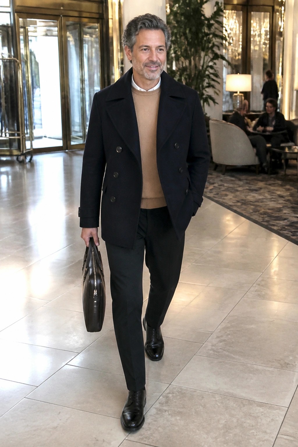 Man in navy peacoat and beige sweater carrying leather briefcase in lobby