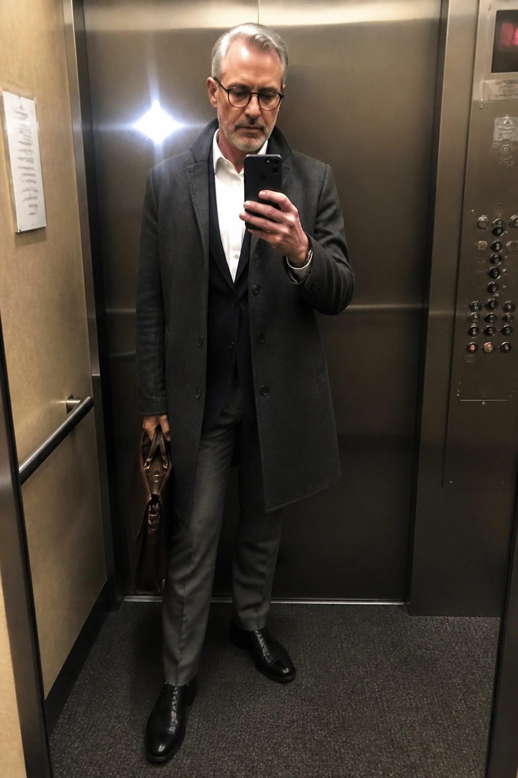 Man in gray overcoat and vest takes selfie in elevator holding briefcase.