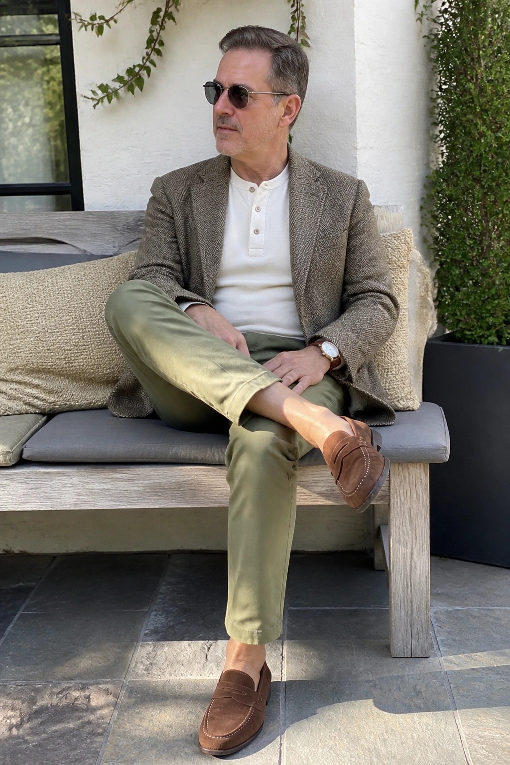 Stylish man in tweed blazer, white henley, and olive pants on outdoor bench.