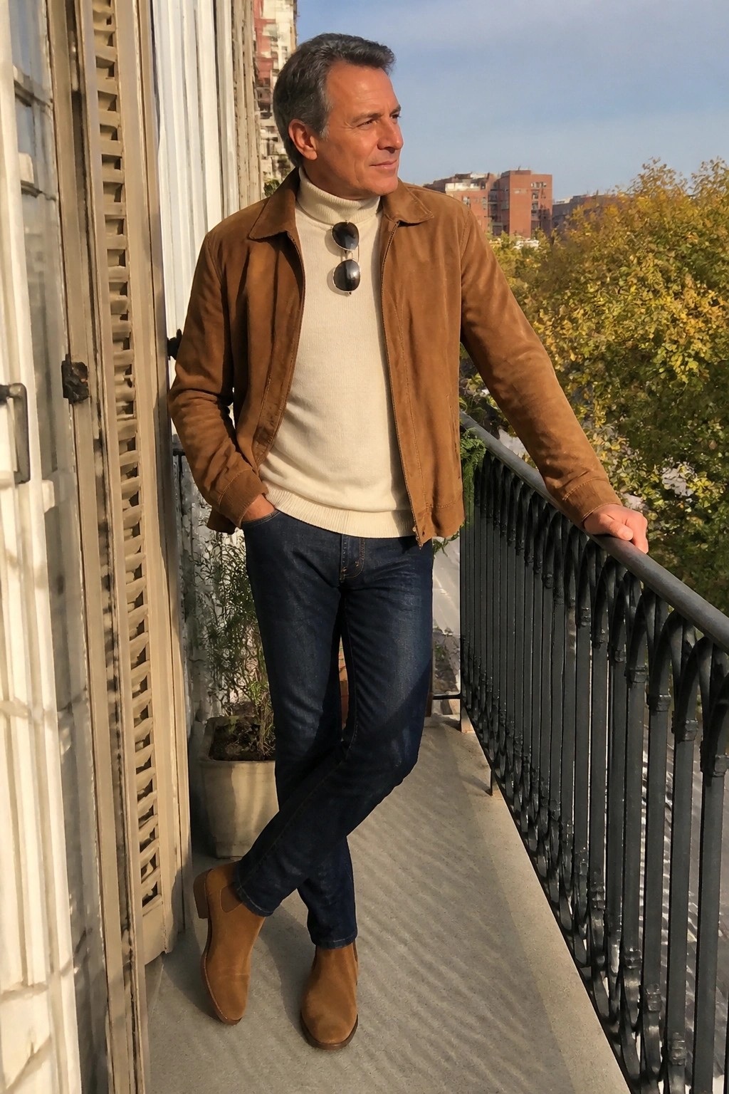 Man in brown suede jacket and white turtleneck leaning on city balcony railing