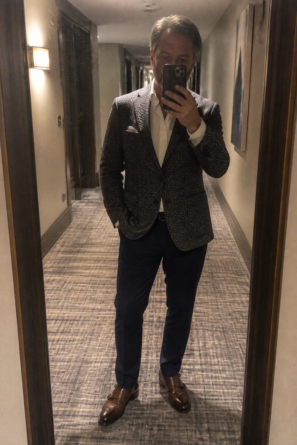 Man in patterned blazer and monk strap shoes takes mirror selfie in hallway