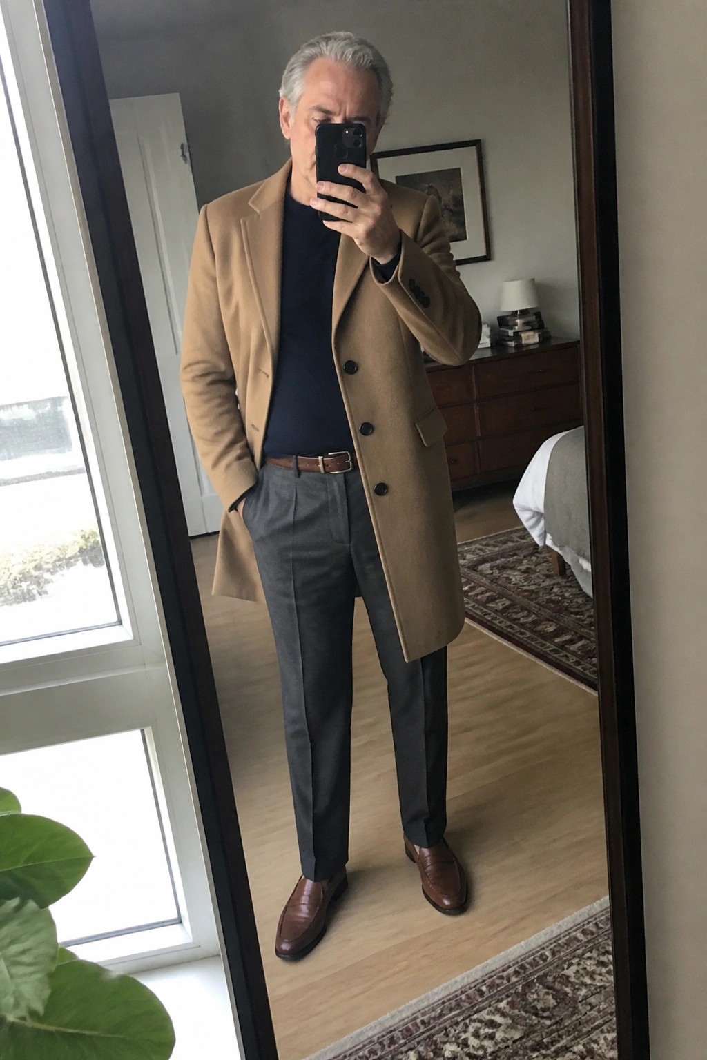 Man in tan overcoat, navy sweater and gray trousers taking mirror selfie