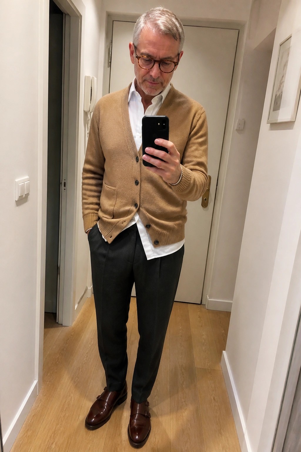 Man in tan cardigan over white shirt with dark trousers taking mirror selfie in hallway