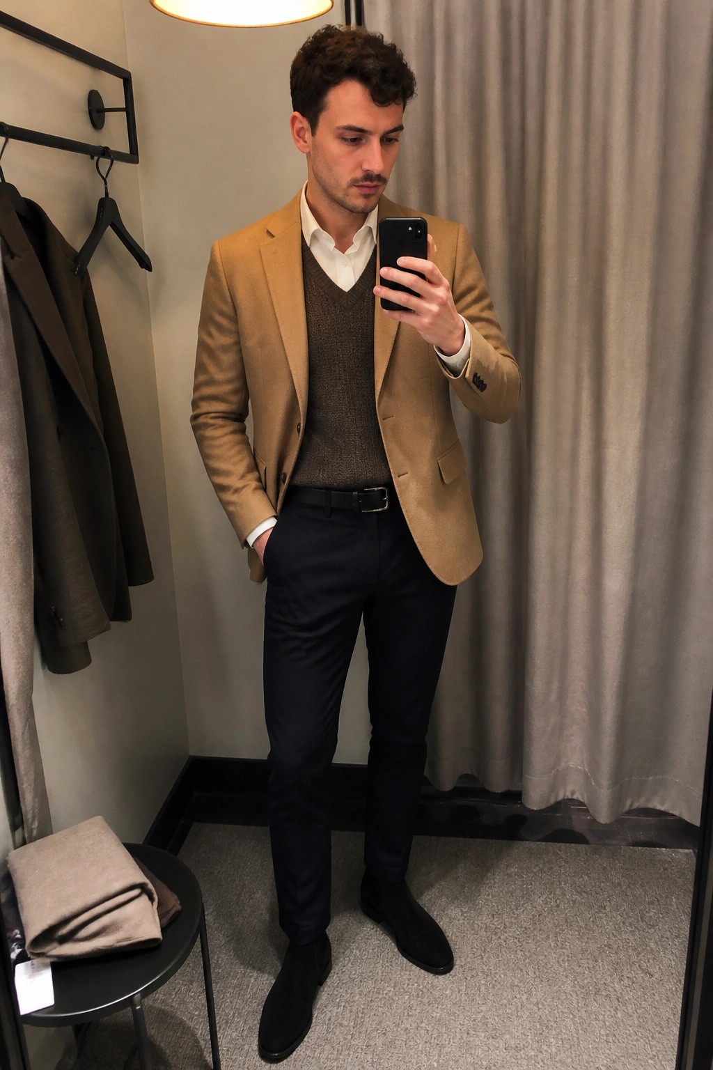 Man in tan blazer and brown knit vest taking mirror selfie in fitting room