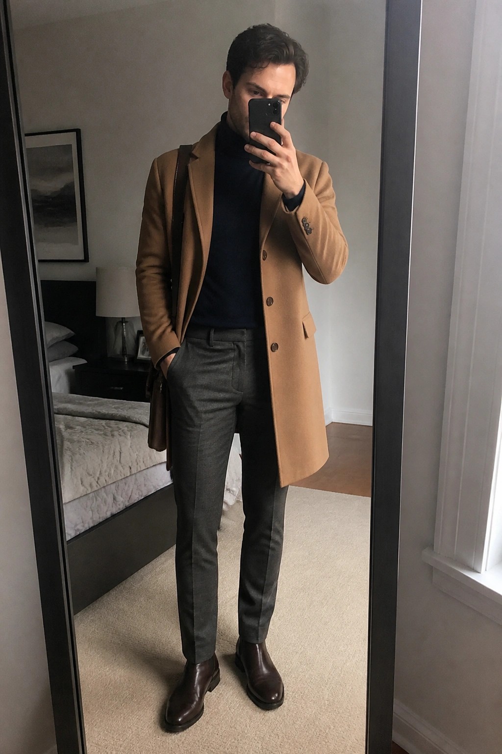 Man in tan overcoat and navy turtleneck taking mirror selfie in bedroom