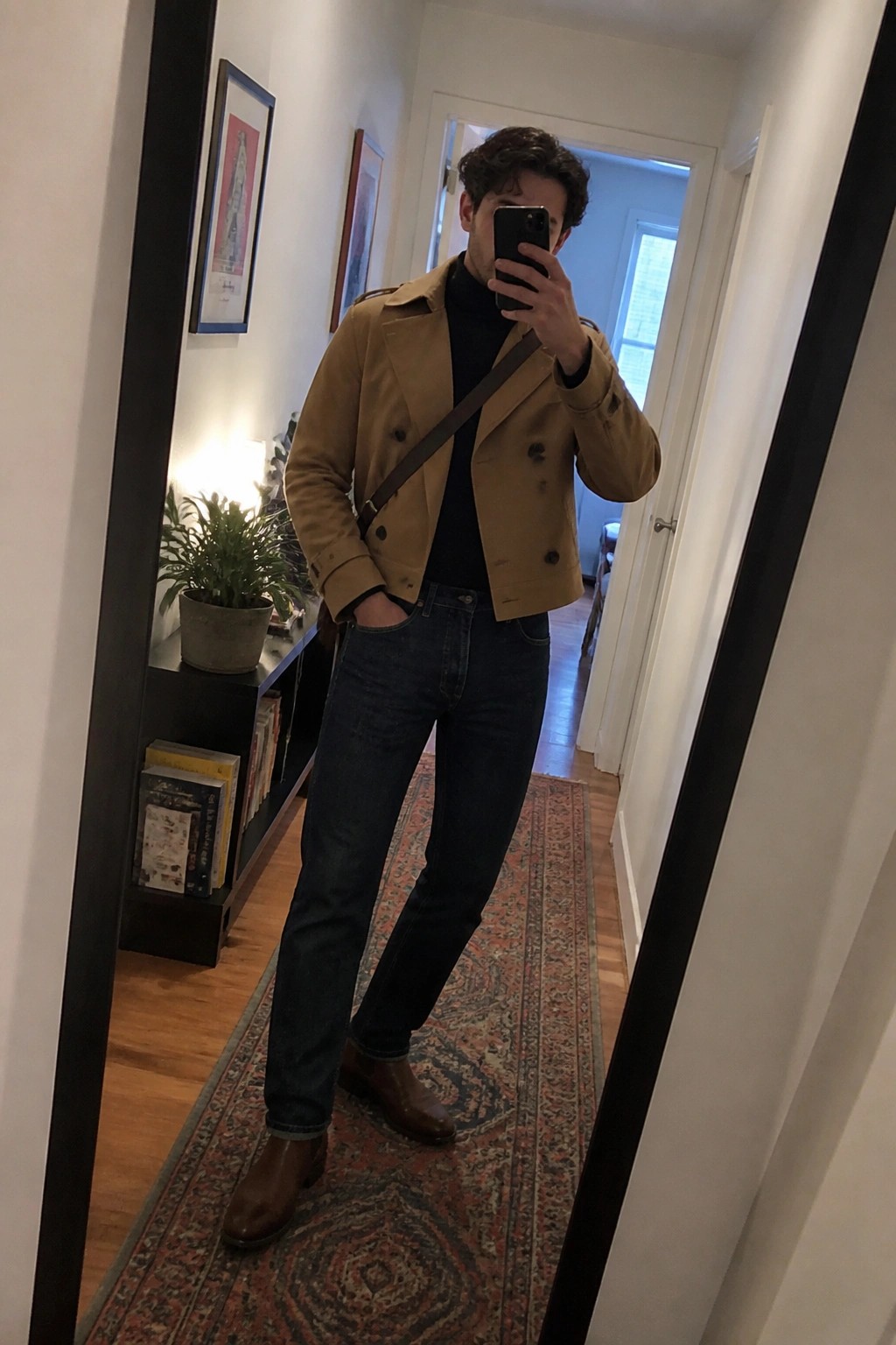 Man in tan jacket and jeans taking mirror selfie in hallway