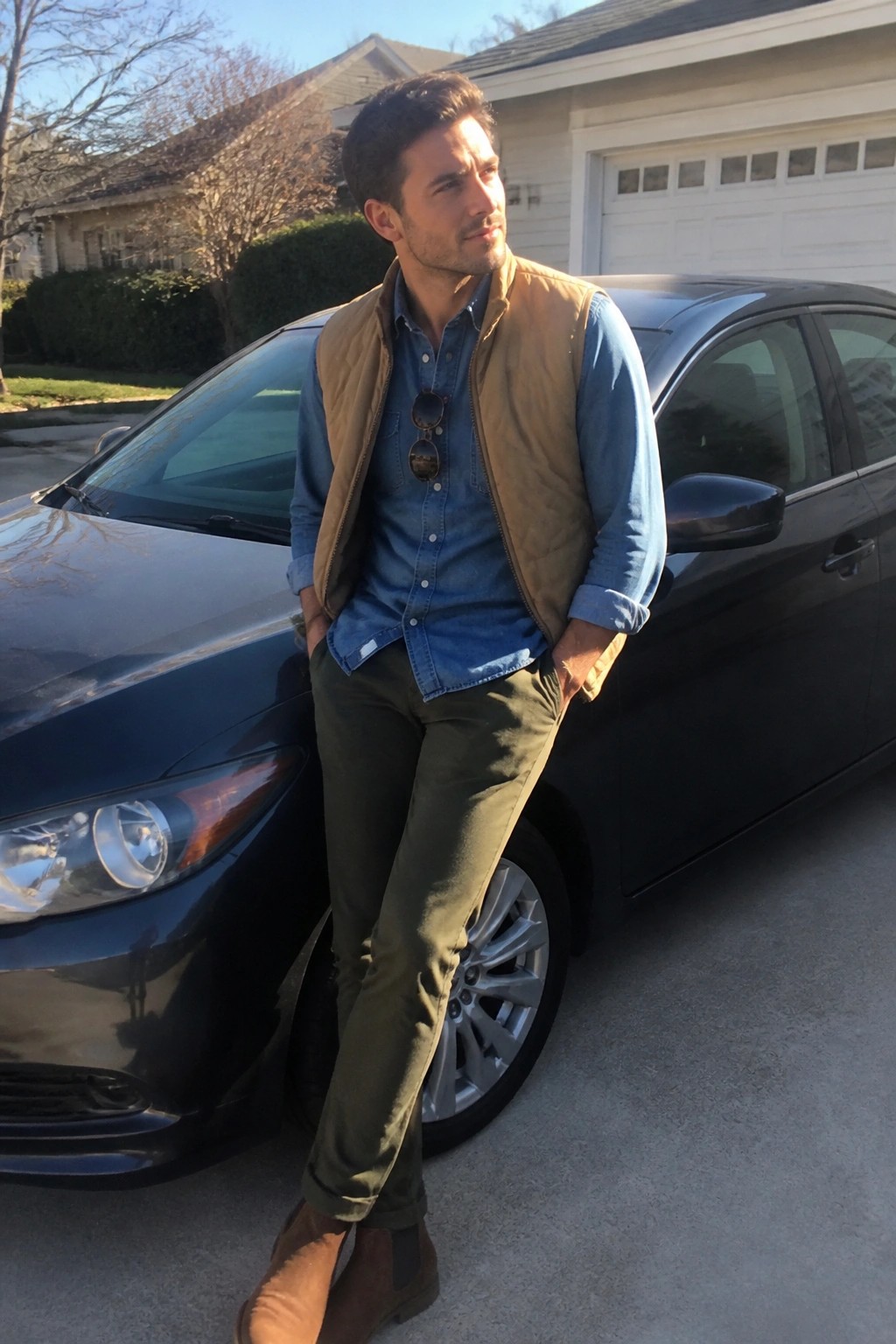 Man in blue denim shirt and tan vest leaning against black car outdoors