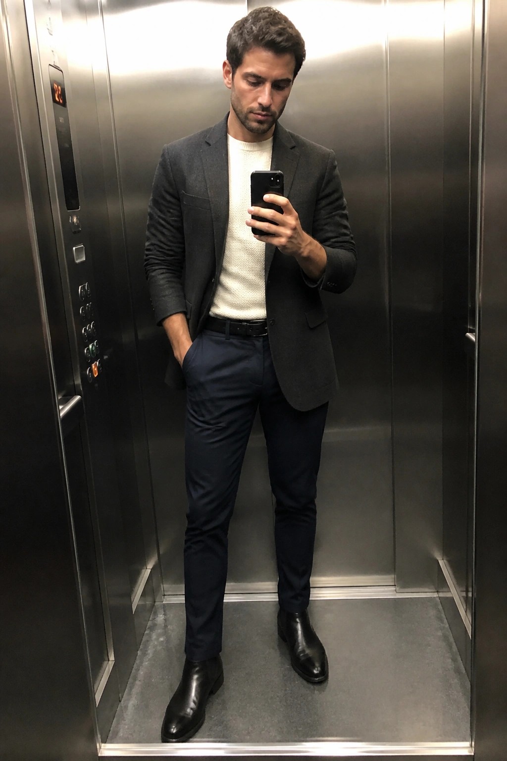 Man in gray blazer over white sweater holds phone in metallic elevator